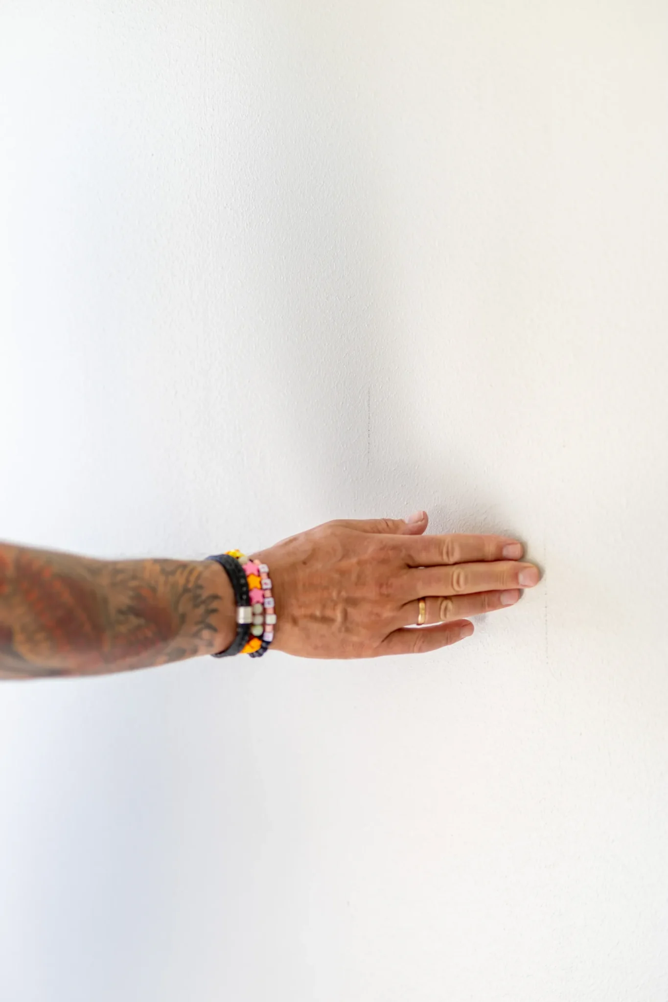 En tatuerad arm med pärlband och ringar rör försiktigt vid en vit vägg, med fingrarna utspridda.