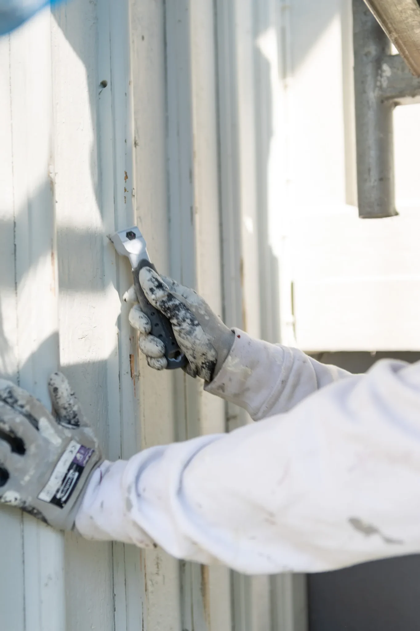 En person som bär handskar och en vit långärmad skjorta skrapar gammal färg från en trävägg eller panel med ett handhållet verktyg.