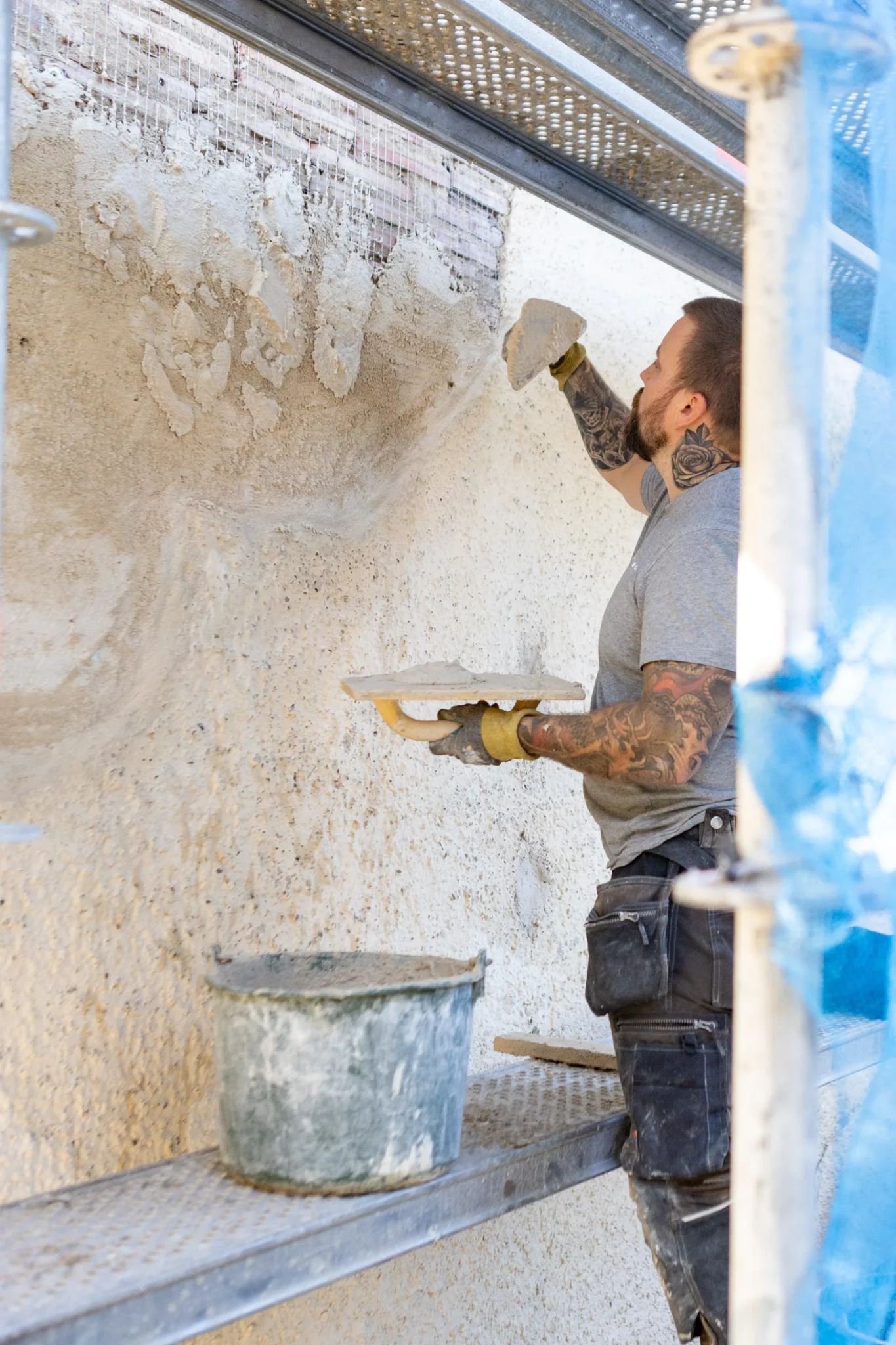 En byggnadsarbetare med tatueringar applicerar gips på en vägg med en murslev medan han står på byggnadsställningar. En hink med gips står i närheten, och arbetaren är fokuserad på att jämna ut ytan.