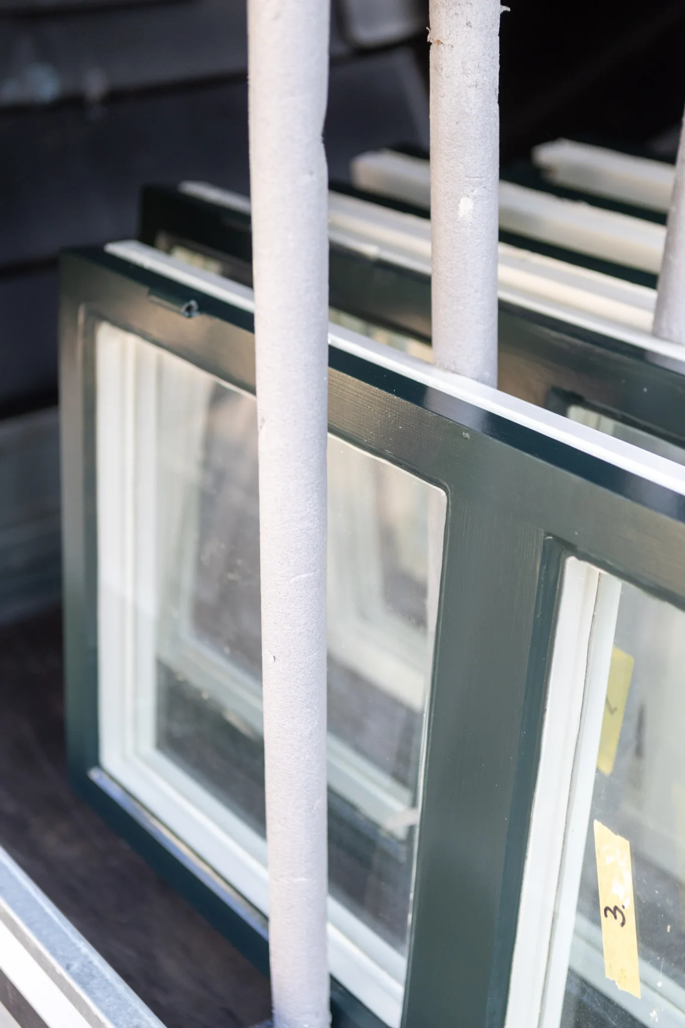Flera fönsterramar med mörkgrön kant och klart glas är staplade vertikalt bakom vadderade metallskenor. Bakgrunden är ur fokus och framhäver fönstren i förgrunden.