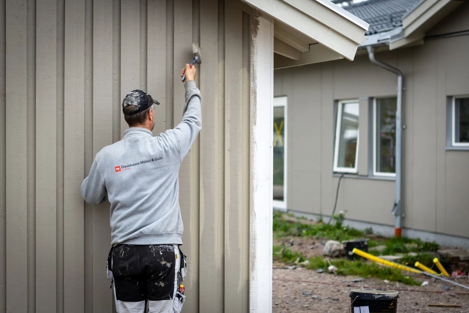 En person som bär en grå jacka målar ytterväggen på ett hus med en pensel. Marken är bar och ett annat hus syns i bakgrunden.