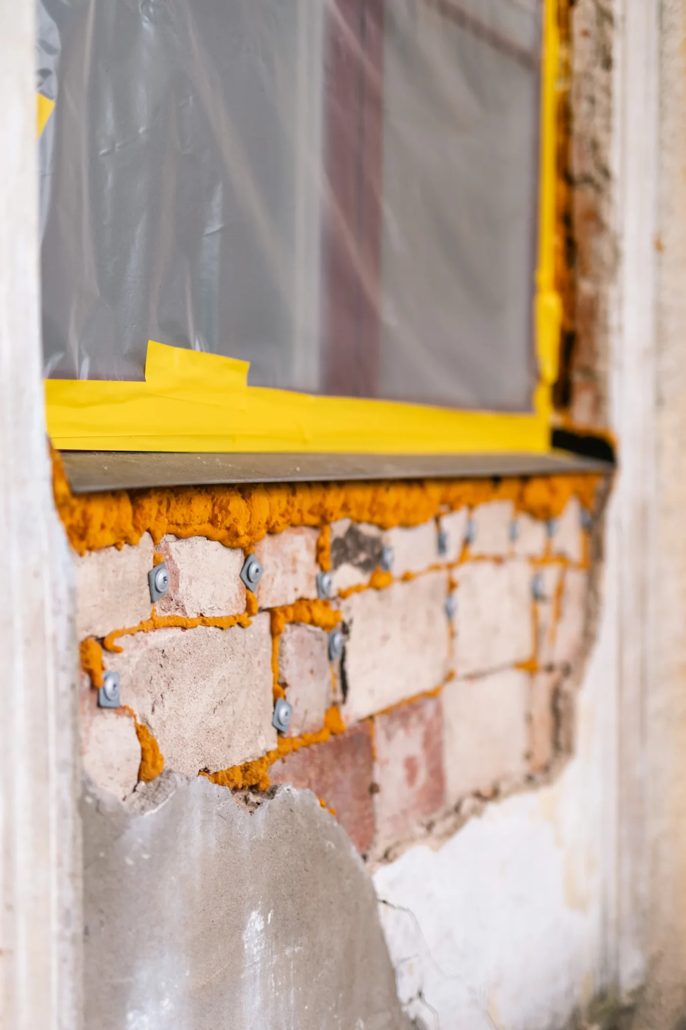 En närbild av en delvis renoverad vägg med exponerade tegelstenar och orange isoleringsskum under ett fönster täckt av plastduk fäst med gul tejp. En del puts saknas i nedre delen av väggen.