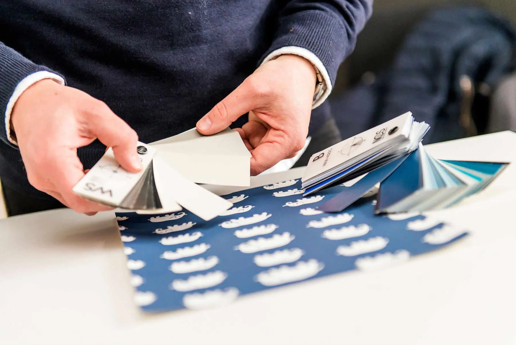 En person håller och undersöker färgprover över ett blått papper med vita kronmönster, och väljer eventuellt färger för design eller dekoration.