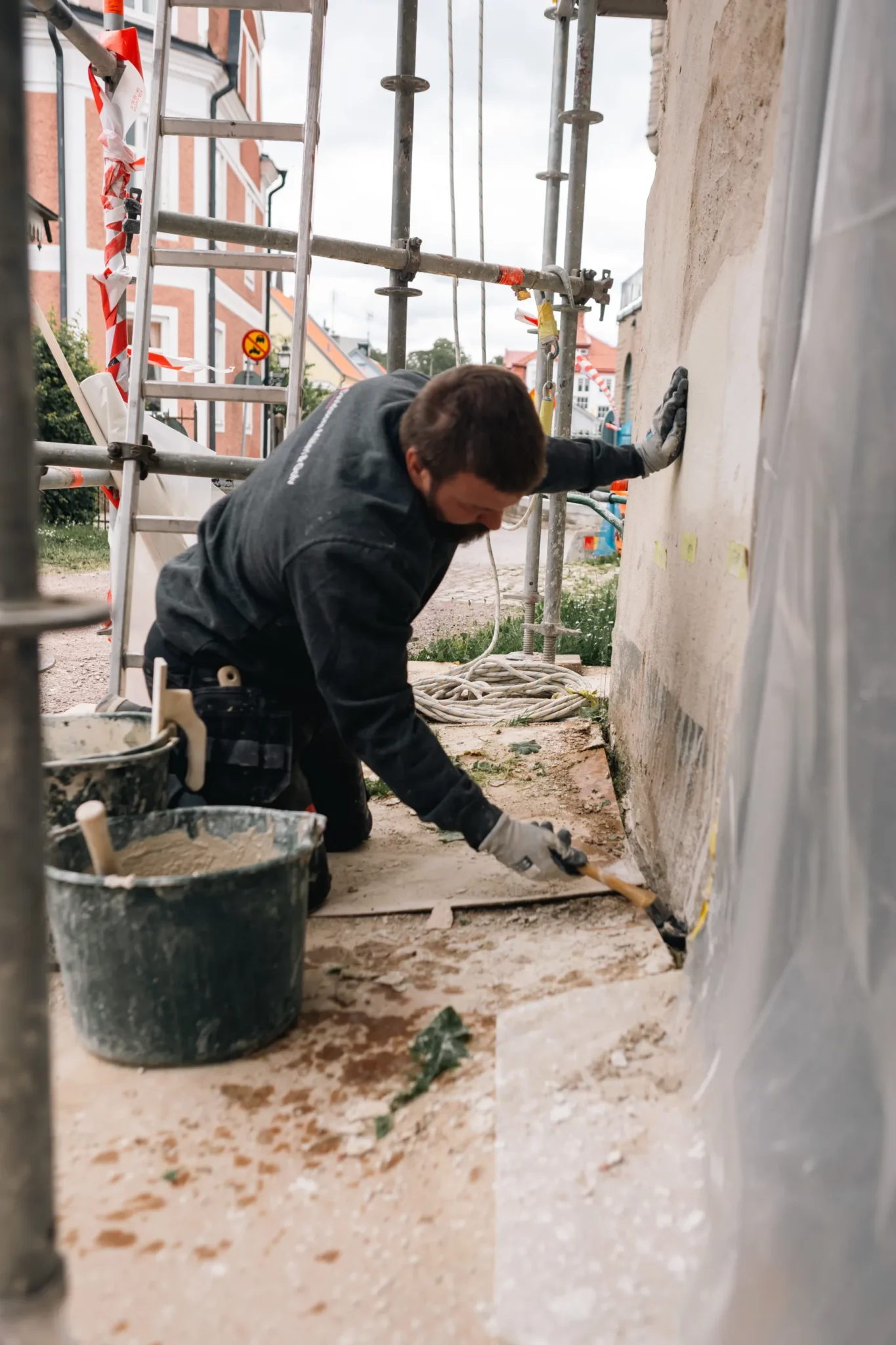 En byggnadsarbetare som bär handskar knäböjer på marken och använder en borste för att applicera material på basen av en byggnadsvägg omgiven av byggnadsställningar och byggmaterial.