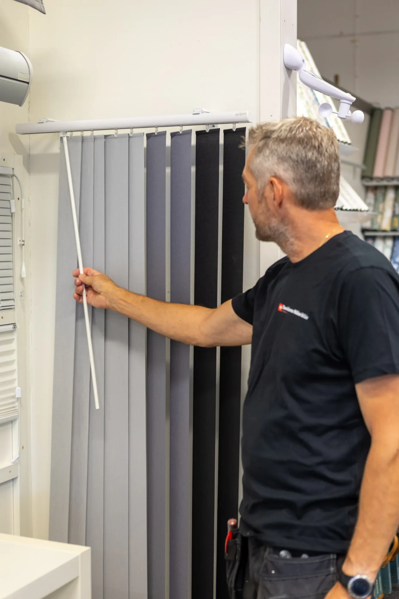 En man med kort hår klädd i en svart t-shirt justerar en uppsättning vertikala persienner med olika grå nyanser med hjälp av en kontrollstav i en butik eller showroom.