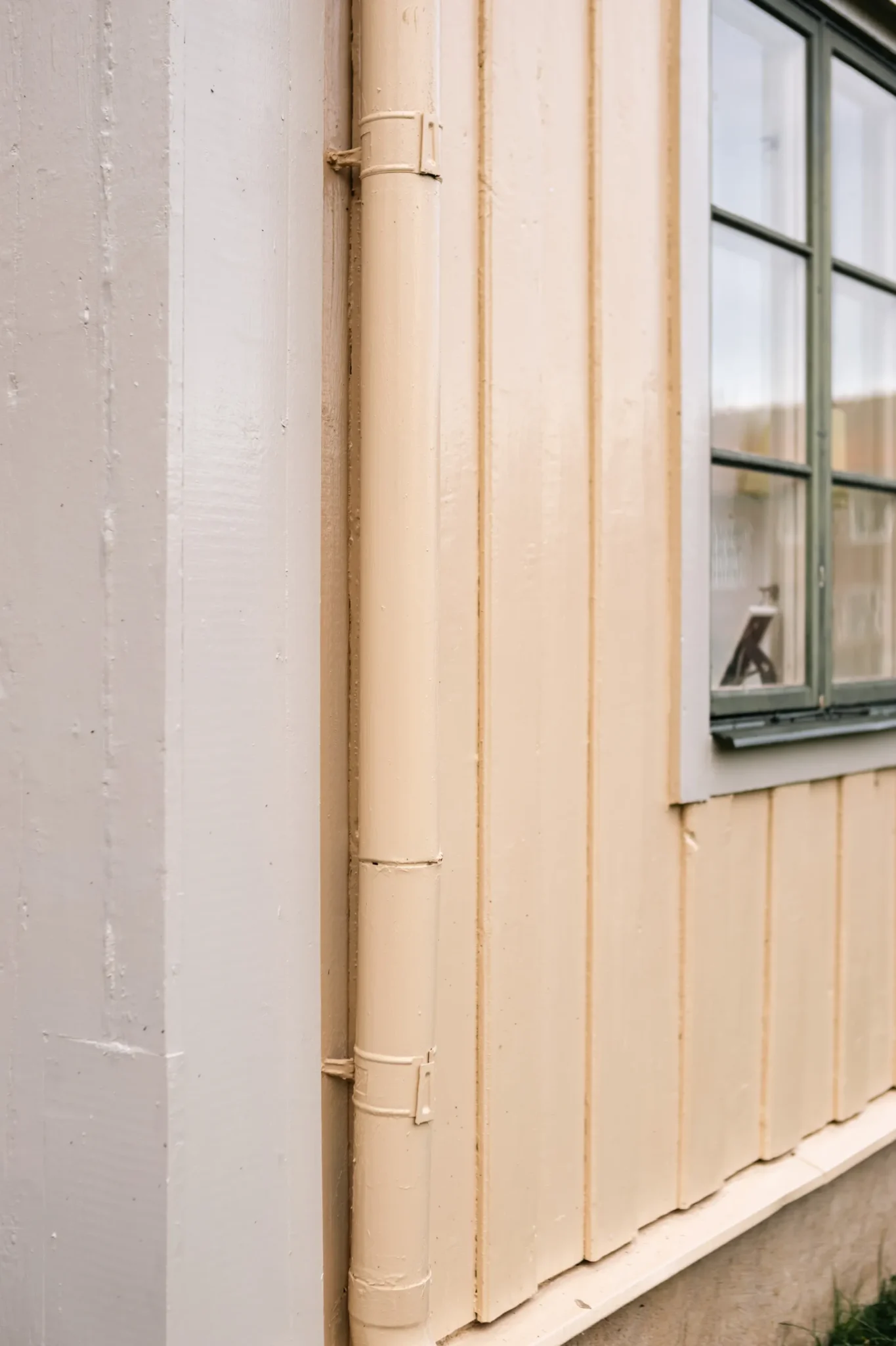 Närbild av hörnet av ett ljust beige trähus med vertikala paneler, ett dräneringsrör och en del av ett fönster med en mörkgrön ram.