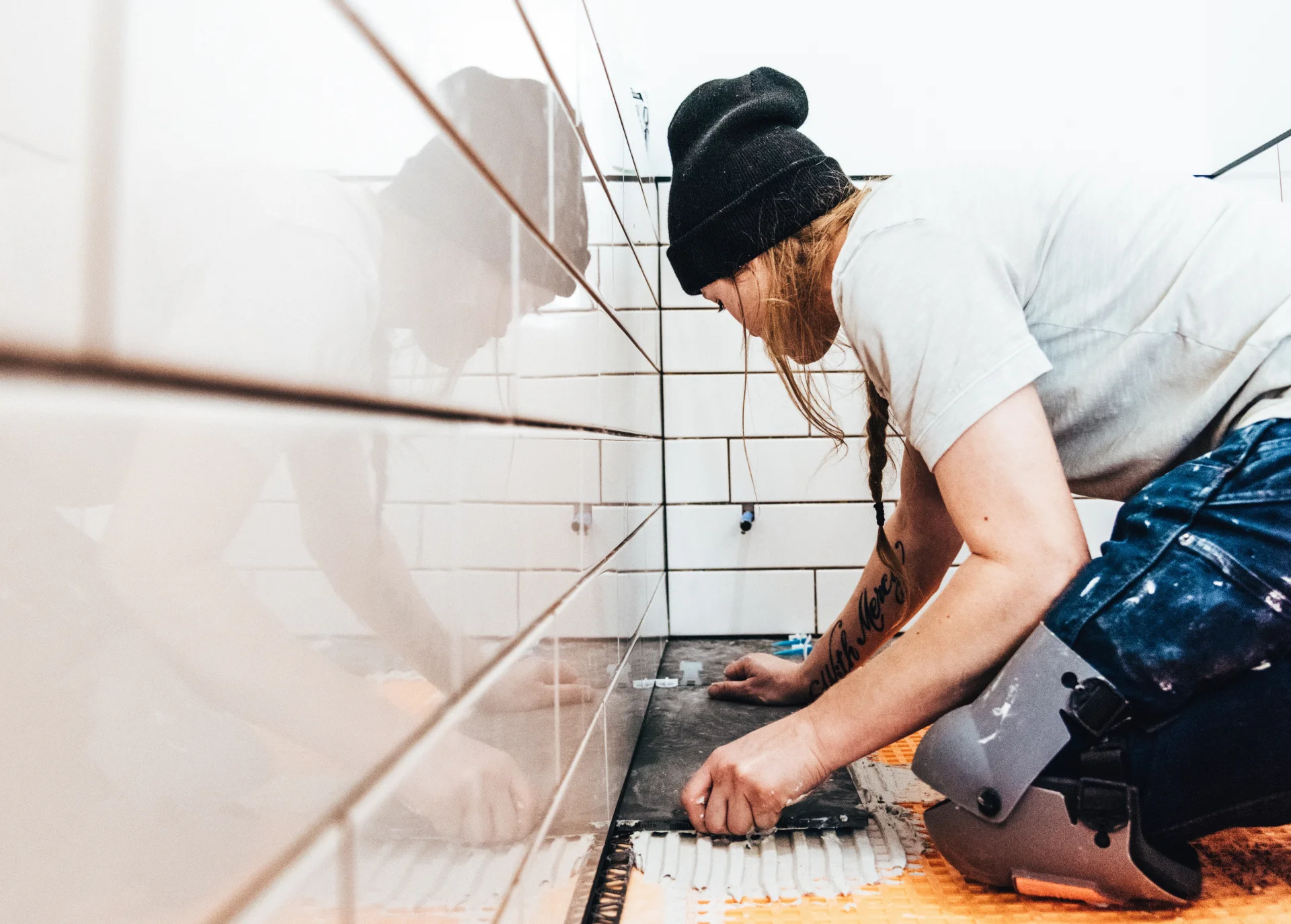 En person som bär mössa och knäskydd sätter vita kakelplattor på en vägg, breder ut lim och lägger en kakel försiktigt. Scenen visar närbilder av deras arbete och koncentration.