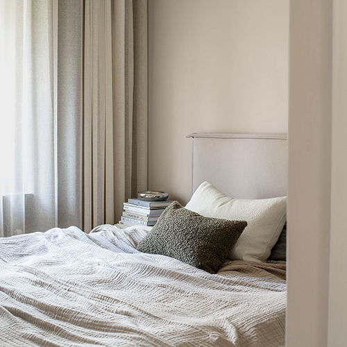 En snyggt bäddad säng med en texturerad grå filt och två kuddar sitter vid ett fönster med ljusa gardiner. På nattduksbordet vilar bokhögar som skapar en lugn och mysig sovrumsatmosfär.