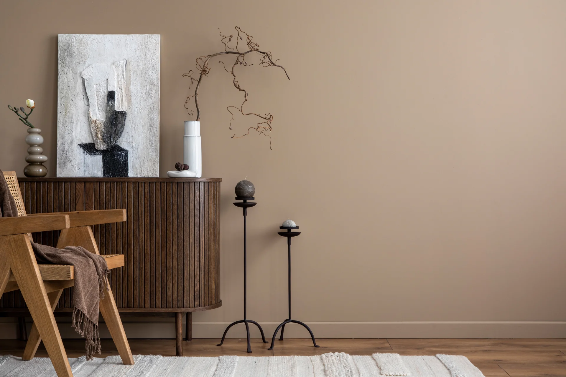Ett minimalistiskt vardagsrum med en trästol, en brun filt, ett texturerat skåp med vaser och abstrakt konst, två smala växtställ och en beige vägg, vilket skapar en lugn och modern atmosfär.