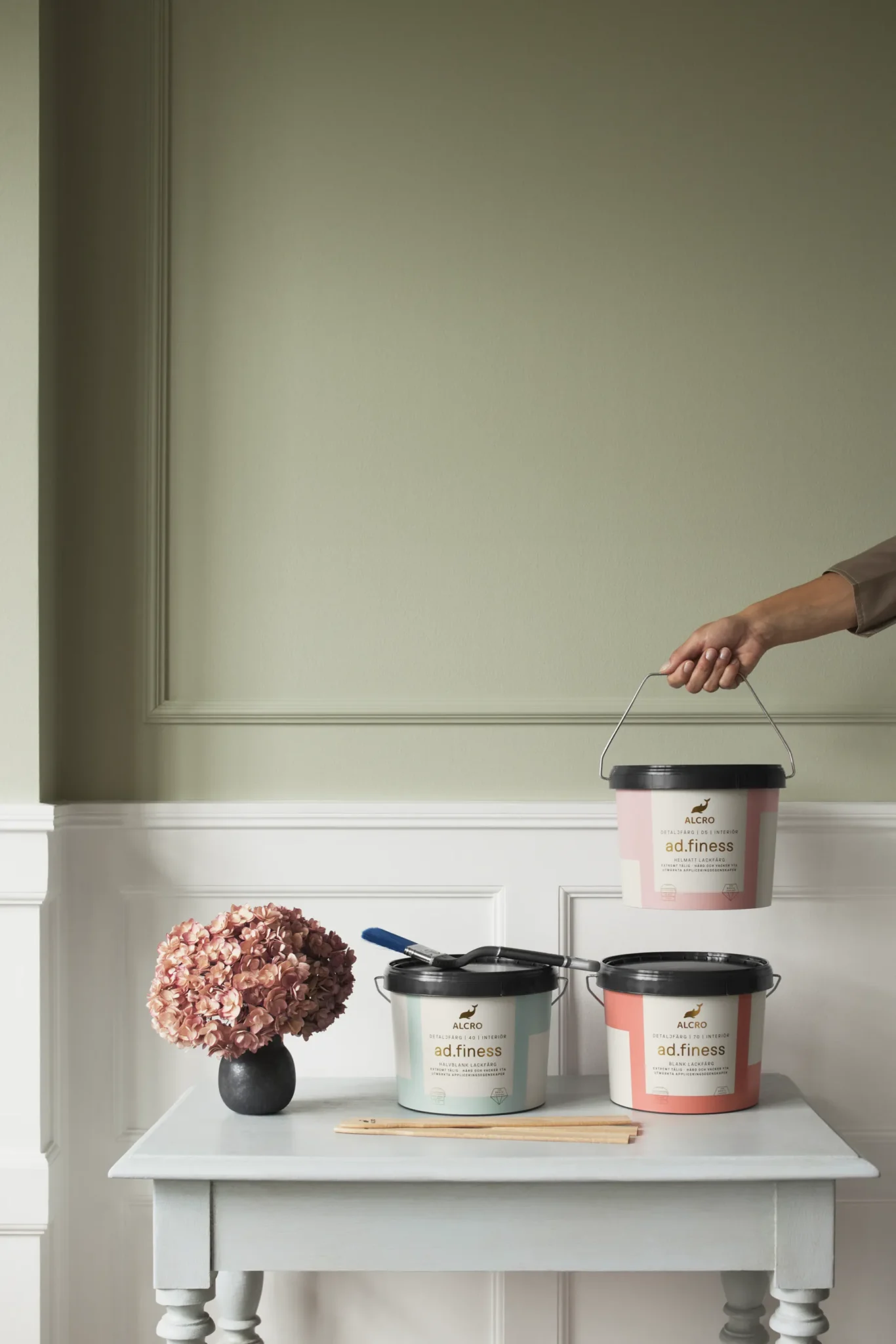 Ett litet bord med två färgburkar, en pensel och en vas med torkade blommor står mot en ljusgrön vägg. En hand håller en tredje färgburk ovanför bordet. Scenen har en minimalistisk och elegant känsla.