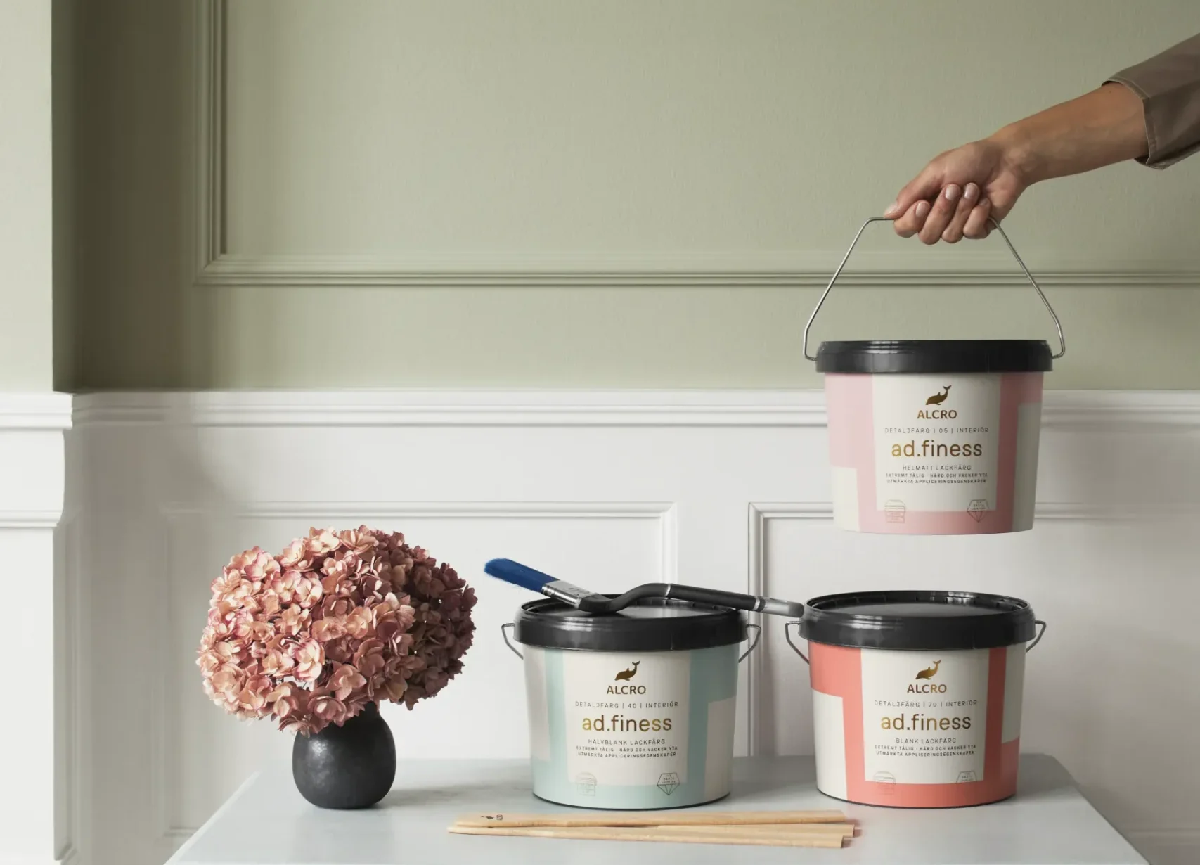 Ett litet bord med två färgburkar, en pensel och en vas med torkade blommor står mot en ljusgrön vägg. En hand håller en tredje färgburk ovanför bordet. Scenen har en minimalistisk och elegant känsla.