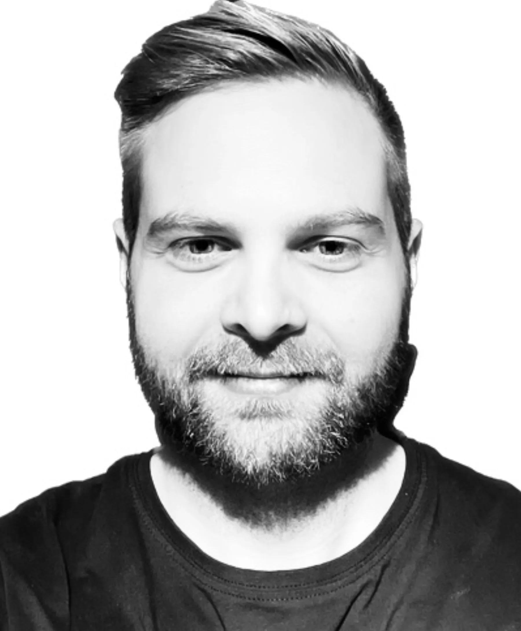 En man med kort, stylat hår och skägg ler lätt mot kameran. Han är klädd i mörk skjorta mot en vit bakgrund. Bilden är i svartvitt.