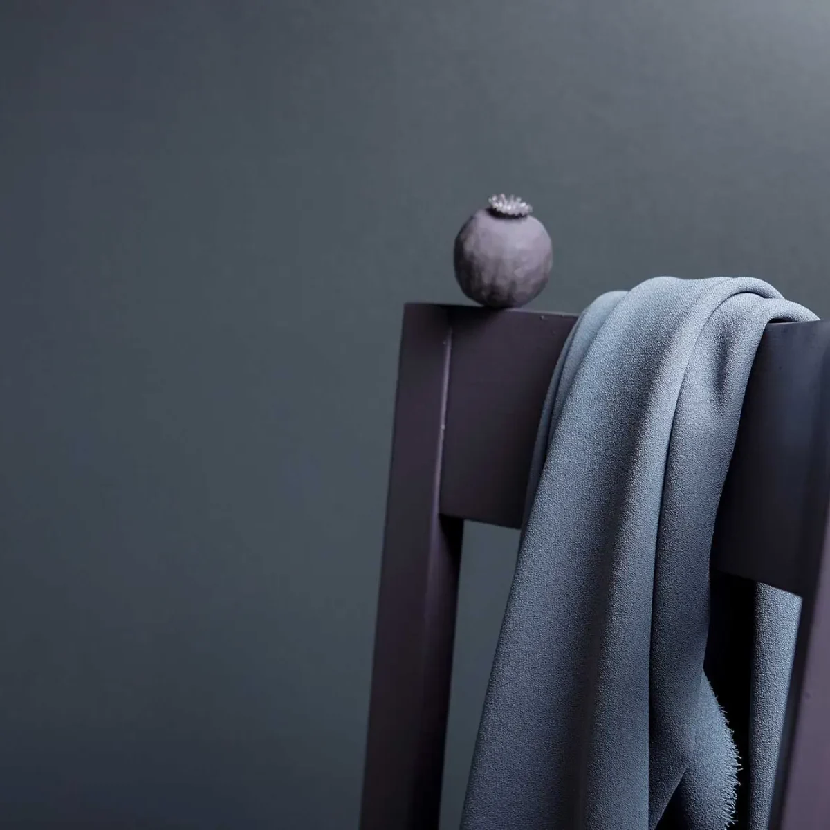 bla-vagg-2000px Ett grått tyg draperat över ryggen på en mörk trästol, med ett litet runt föremål, eventuellt en torkad fröskida, placerad ovanpå stolen mot en vanlig mörk bakgrund.