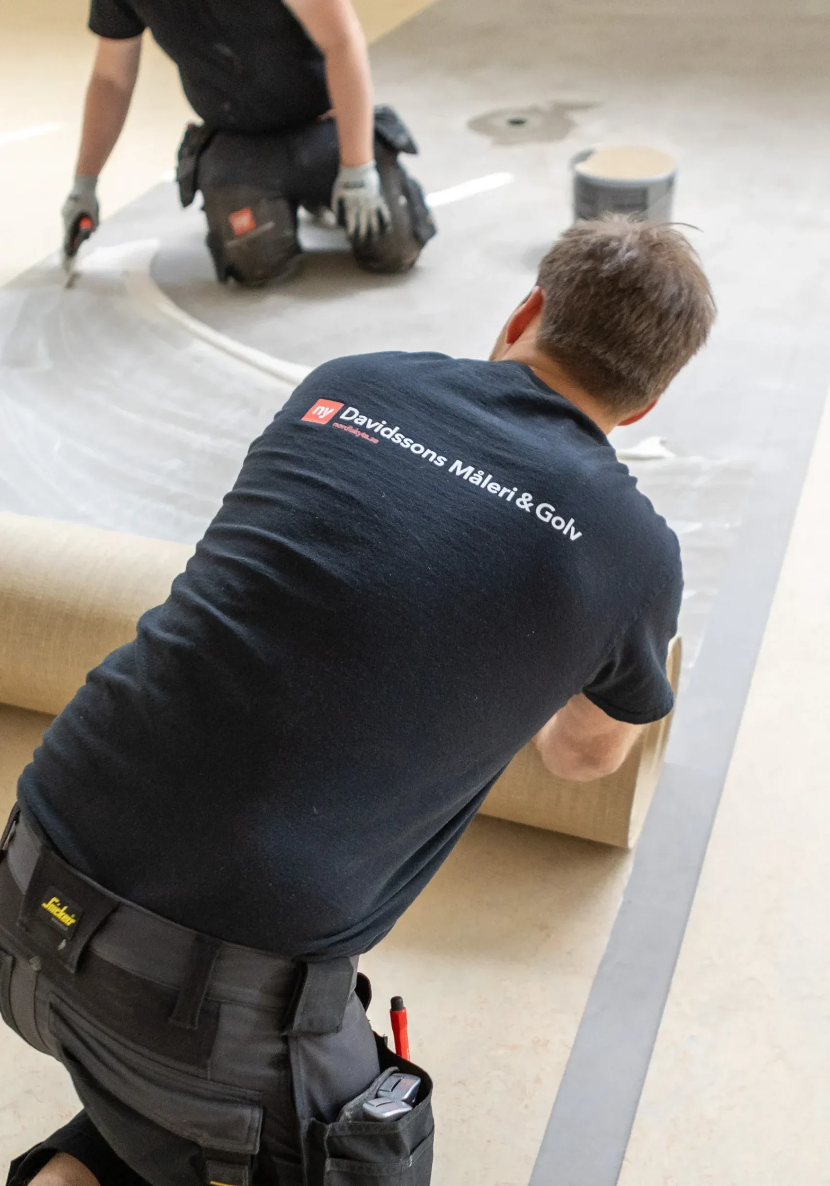 Två arbetare lägger en rulle golvbeläggning på lim på marken. Den ena arbetaren rullar ut materialet medan den andra slätar ut det. Båda är klädda i svarta skjortor och arbetsbyxor.