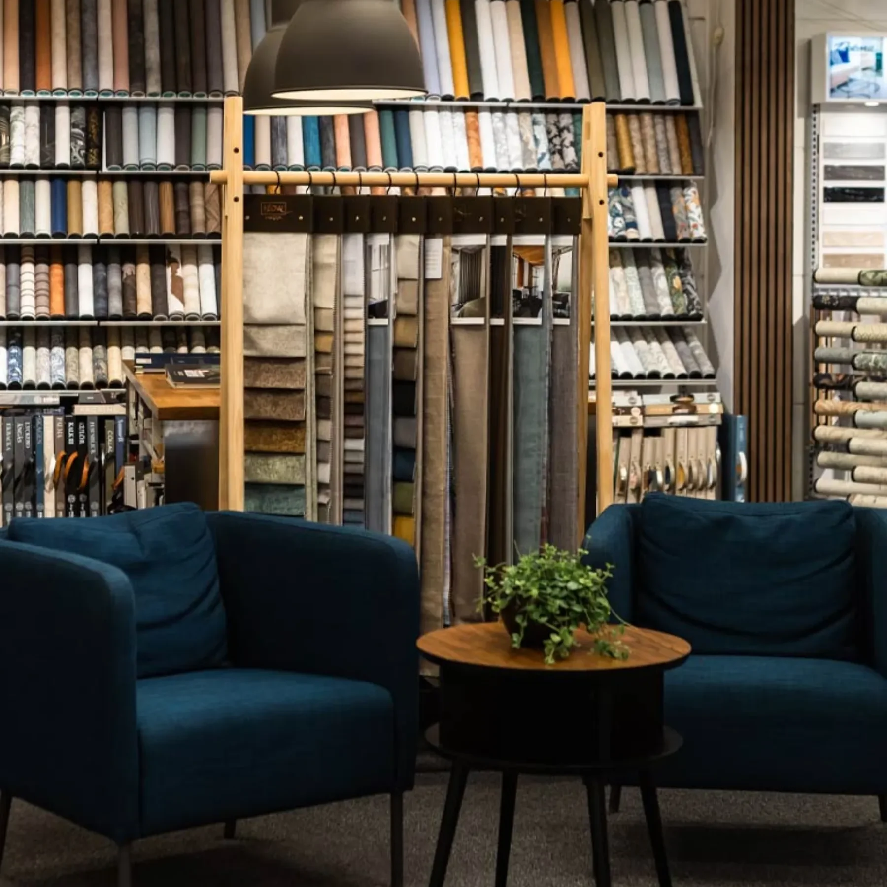 Två mörkblå fåtöljer och ett litet runt bord med en krukväxt står framför skyltställ fyllda med tygrullar och mattprover i ett modernt välupplyst showroom.